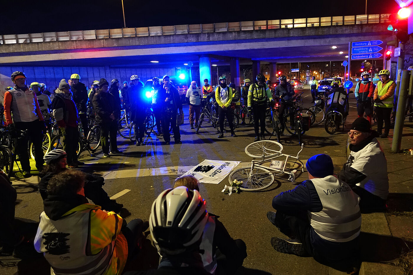 Menschen sitzen bei einer Mahnwache um ein weißes Fahrrad auf der Straße