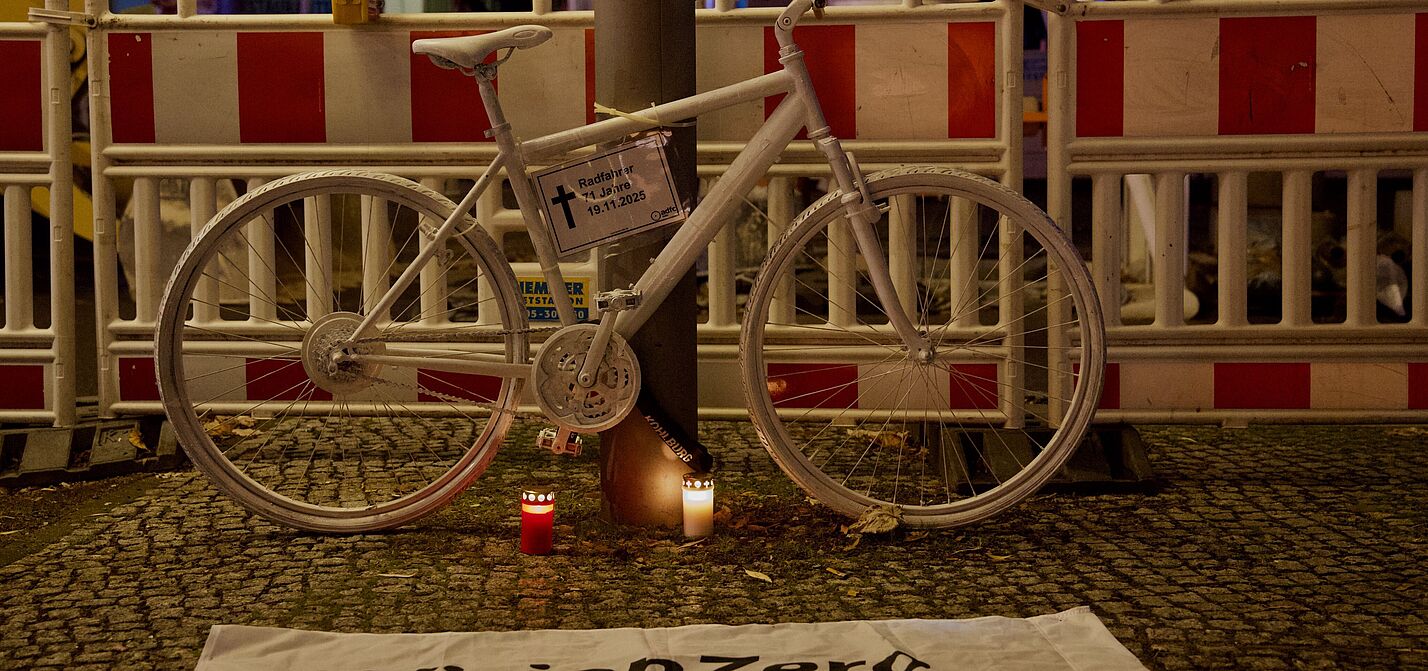 Ein weißes Geisterrad an einem Laternenphal davor eine weiße Vision Zero Fahne