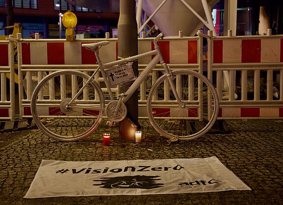 Ein weißes Geisterrad an einem Laternenphal davor eine weiße Vision Zero Fahne