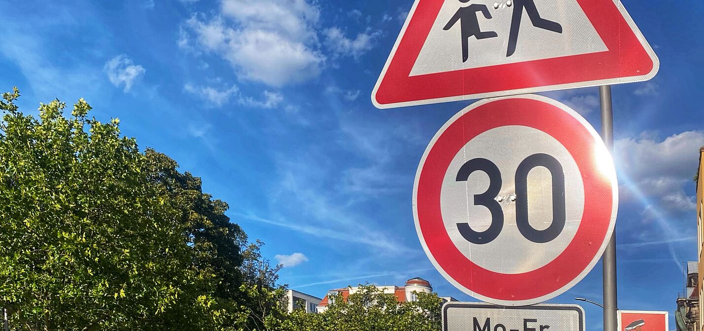 Tempo 30 auf dem Schulweg an einer Hauptverkehrsstraße: Grunewaldstraße in Berlin Schöneberg Auf dem Foto sind zwei Verkehrsschilder vor blauem Himmel zu sehen: Tempo 30 und Kinder. Rechts liegt eine Häuserzeile, auf der linken Seite ist die Straße durch Bäume begrünt.