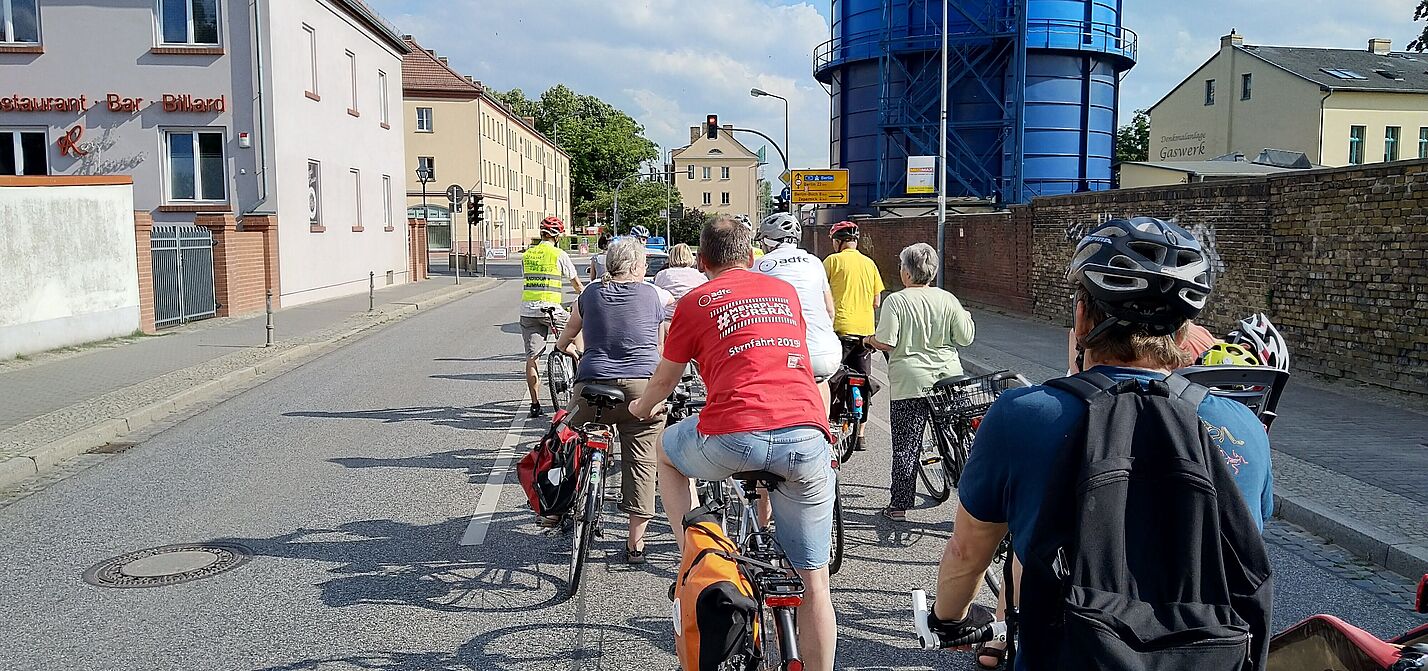 Das Bild zeigt eine Gruppe von Radfahrern auf einer städtischen Straße. Sie stehen hintereinander auf ihren Fahrrädern und tragen teils Helme sowie Rucksäcke. Links sind Wohnhäuser, rechts eine Backsteinmauer und der Bernauer Gaskessel (ein großes, blaues, rundes Industriegebäude) zu sehen. Das Wetter ist sonnig und freundlich. 