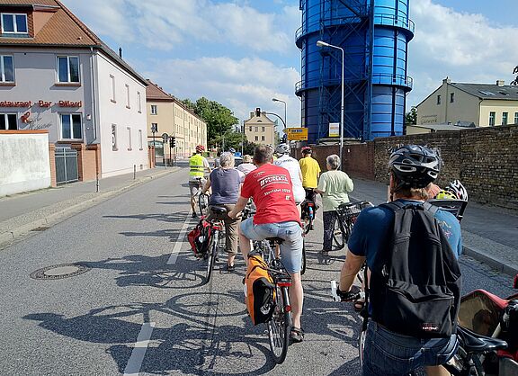 Das Bild zeigt eine Gruppe von Radfahrern auf einer städtischen Straße. Sie stehen hintereinander auf ihren Fahrrädern und tragen teils Helme sowie Rucksäcke. Links sind Wohnhäuser, rechts eine Backsteinmauer und der Bernauer Gaskessel (ein großes, blaues, rundes Industriegebäude) zu sehen. Das Wetter ist sonnig und freundlich. 